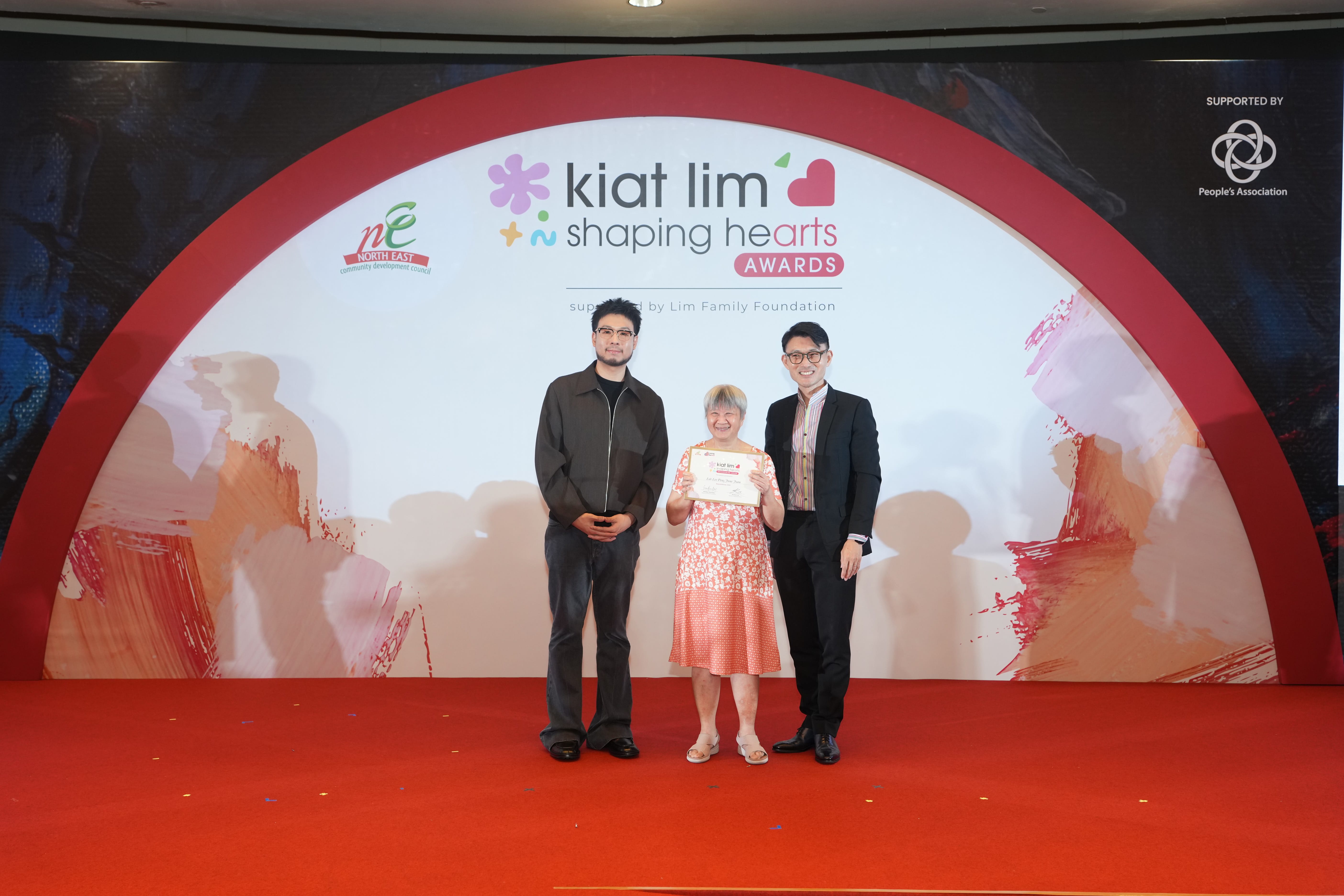 Award recipient standing on stage with the presenter, holding their certificate and posing for a group photo during the award ceremony, with a backdrop reading ‘Kiat Lim-Shaping Hearts Awards'.